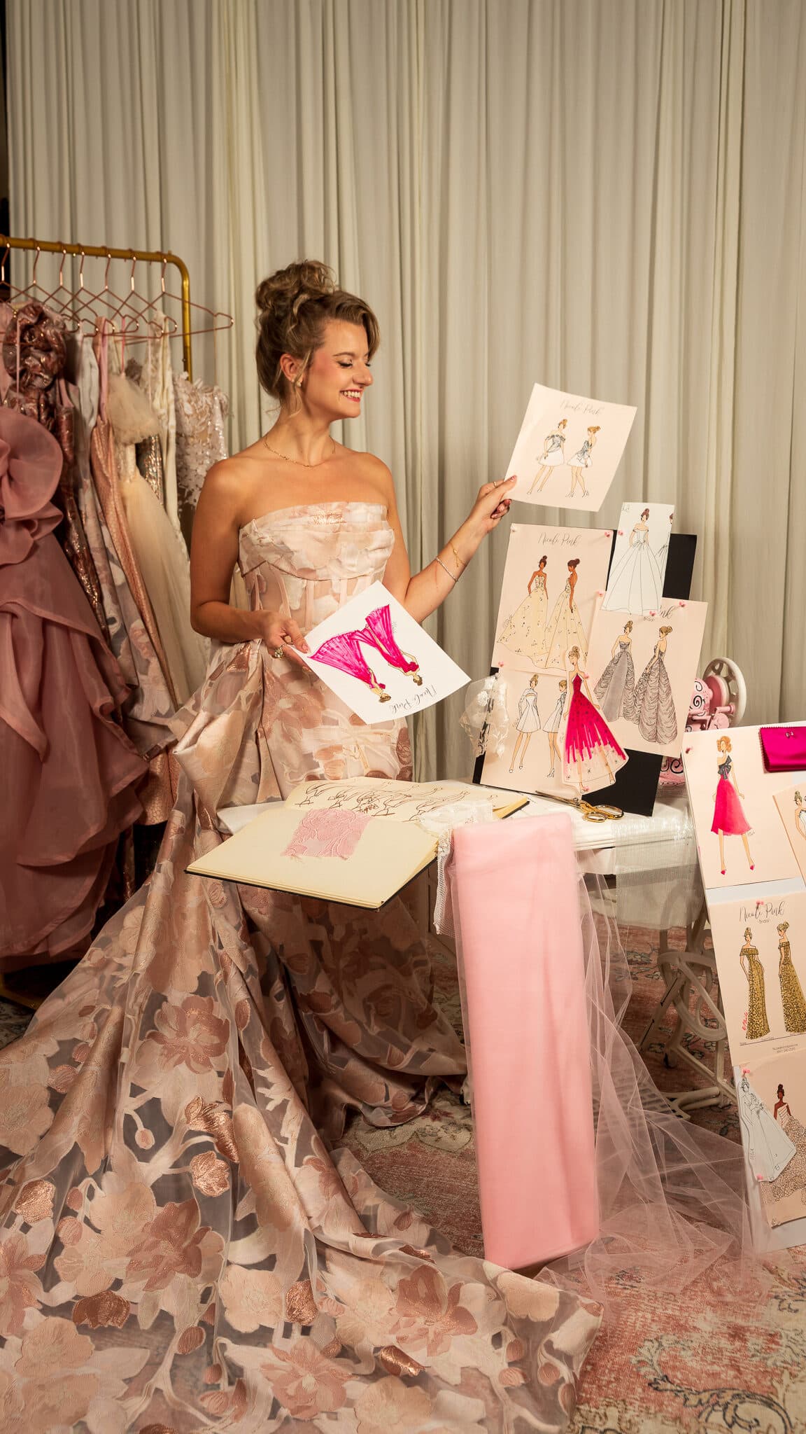 Nicole Pink in a blush floral gown, holding a fashion illustration beside a board of her sketches