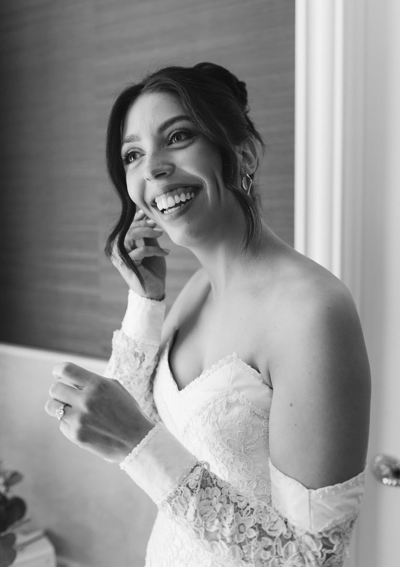 Bride laughing by a window in an off the shoulder lace gown, in black and white
