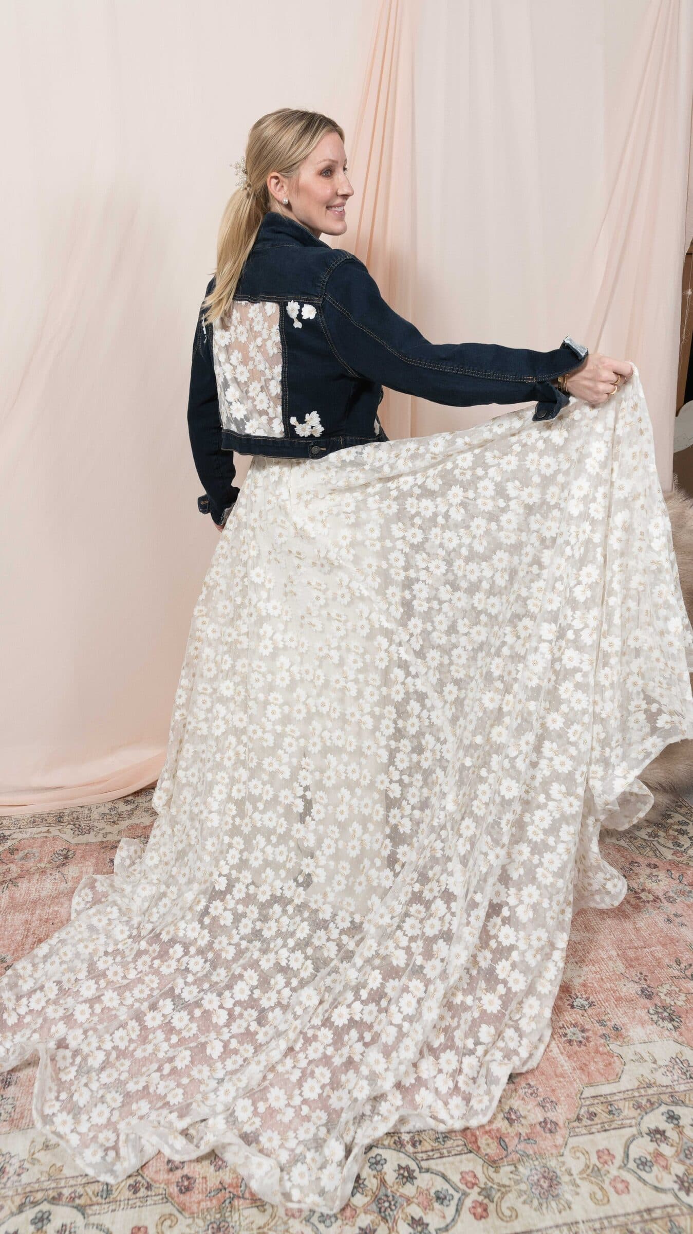 Bride in an embellished denim jacket holding out the skirt of a white floral lace gown